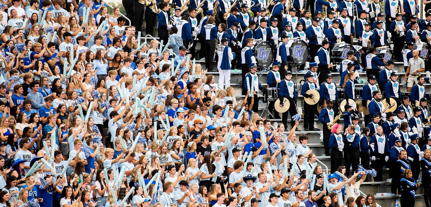 A large crowd in blue and white attire cheers enthusiastically at a stadium, with a marching band performing on the steps.